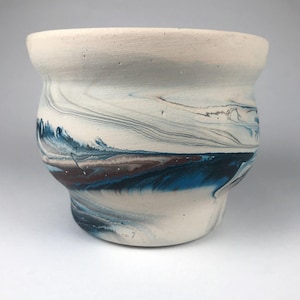 May include: A white ceramic planter with a marbled design in shades of blue, brown, and gray. The planter has a wide rim and a rounded body, perfect for displaying plants. The interior is white.