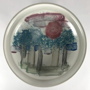May include: A white ceramic plate with a hand-painted design of trees, a red sun, and blue and white abstract shapes.