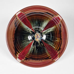 May include: A handmade ceramic bowl with a unique, colorful design. The bowl features a radial pattern with red, black, and white stripes, and a glossy finish. The center has a swirling, multi-colored design. The rim is a reddish-brown color.