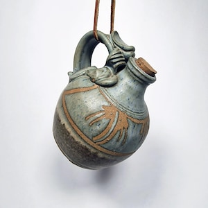 Può includere: Piccolo boccale in ceramica blu-grigio con tappo in sughero e cordino in pelle marrone. Il boccale presenta un design decorativo in un colore marrone più chiaro. L'impugnatura è curva e ha un elemento decorativo nella parte superiore. Il boccale è sospeso su sfondo bianco.