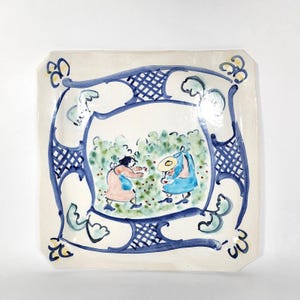 Puede incluir: Plato cuadrado de cerámica con diseño pintado a mano. El plato presenta un borde azul y blanco con una ilustración caprichosa de dos figuras en un jardín. Las figuras llevan sombreros y sostienen objetos. El plato tiene una forma ligeramente irregular.