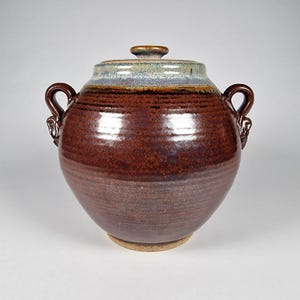 Może przedstawiać: Ręcznie wykonany ceramiczny słoik z pokrywką. Słoik jest w bogatym brązowym kolorze z jaśniejszą brązową podstawą i niebiesko-szarym pasmem w pobliżu góry. Ma dwa uchwyty i małe pokrętło na pokrywce.