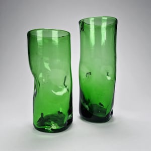 May include: Two tall, cylindrical drinking glasses made of translucent green glass. Each vessel features an irregular, organic shape with subtle indentations along the sides and a thick, slightly uneven base, creating a hand-blown appearance.