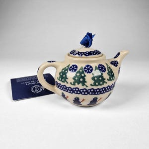 Puede incluir: Pequeña tetera de cerámica color crema con un diseño invernal azul y verde. El diseño incluye árboles de Navidad, copos de nieve y muñecos de nieve. Una cinta azul está atada a la tapa. Una tarjeta azul está al lado de la tetera.