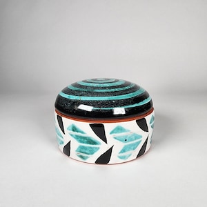 May include: A round ceramic trinket box with a lid. The lid is black with turquoise and black concentric circles. The box has a white base with turquoise and black leaf-like patterns. The rim is brown.