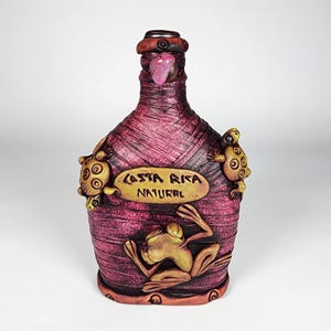 May include: A maroon-coloured bottle with a textured surface, decorated with turtle and frog figurines. The bottle has a label reading "COSTA RICA NATURAL". The top of the bottle features a pink stone and a brown cap.