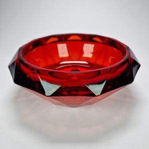 Pode incluir: Um cinzeiro de vidro facetado vermelho rubi. O cinzeiro tem um design geométrico com ângulos agudos e uma tigela circular. O vidro é transparente, permitindo a passagem da luz, e as bordas são escuras.