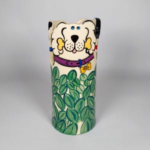May include: A whimsical ceramic vase featuring a cartoon dog design. The vase has a cream-colored base with a dog's face at the top, holding a bone. The dog wears a purple collar with a dog tag. Green leaves decorate the lower portion of the vase.