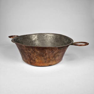 May include: A rustic copper bowl with two circular handles. The bowl has a hammered texture and a dark, aged patina. The interior is a lighter, silvery colour. The bowl is likely used for cooking or serving.