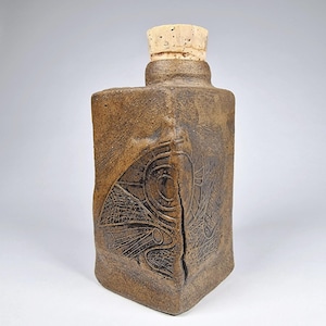 Puede incluir: Botella de cerámica rectangular marrón con tapón de corcho. La botella presenta un diseño grabado en la parte delantera, con una grieta en el centro. El corcho es de color beige claro.