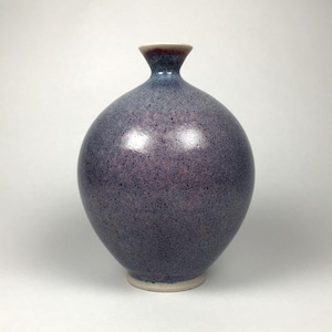 May include: A speckled, spherical vase with a narrow neck and a flared rim. The vase has a gradient of colors, transitioning from a deep purple at the base to a lighter blue at the neck, with a reddish-brown rim. The base is a light cream color.