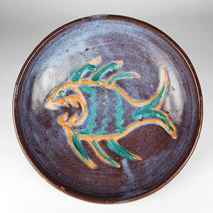 May include: A round ceramic plate with a dark brown and blue glazed finish. The center features a hand-painted fish design in vibrant orange and teal colors, with wavy lines on the body and sharp fins, creating a textured and glossy appearance.