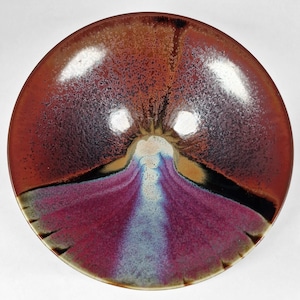 May include: A round ceramic bowl featuring a glossy glaze with a vibrant color gradient. The interior displays a deep reddish-brown rim that transitions into a dark purple and bright magenta center, accented by a streak of light blue and white.