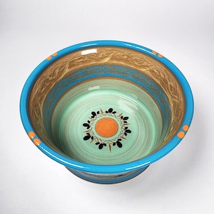 May include: A ceramic bowl with a turquoise rim and a light blue interior. The bowl features a decorative pattern of brown, blue, and orange concentric circles. The center of the bowl has a light green background with an orange circle and black leaf-like shapes.