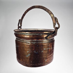May include: A small, antique copper bucket with a curved handle. The bucket has a cylindrical body with a slightly flared rim and a hammered texture. The handle is attached with decorative brackets.