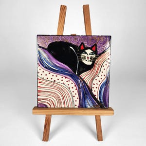 Könnte beinhalten: Ein quadratisches Fliesenbild mit einer schwarzen Katze mit rot gefärbten Ohren vor einem violetten Hintergrund mit goldenen Spiralmustern. Im Vordergrund befinden sich abstrakte, wellenförmige Linien in Blau, Rot und Weiß mit kleinen roten Punkten.