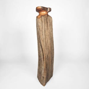 May include: A tall, rustic wooden sculpture with a natural, weathered appearance. The sculpture features a rectangular body with a flared top section in a contrasting copper color. The wood grain is visible, adding texture and character.