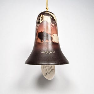 Op de afbeelding: Een klokvormige hanglamp met een bruine basis en een crèmekleurige gloeilamp. De lamp heeft een geschilderd ontwerp van bizons en een landschap. De tekst "Blows, Listen, It speaks" staat op de lamp.