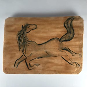May include: A rectangular, tan-colored piece of art featuring a black ink drawing of a running horse. The horse is depicted in a dynamic pose, with its legs extended and tail flowing. The artwork has a textured appearance.