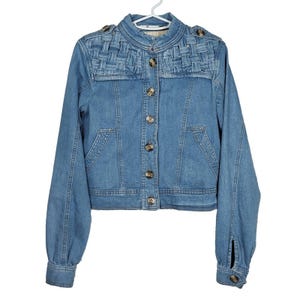 May include: A light blue denim jacket with a woven detail on the shoulders and a button closure. The jacket has long sleeves and a cropped length.