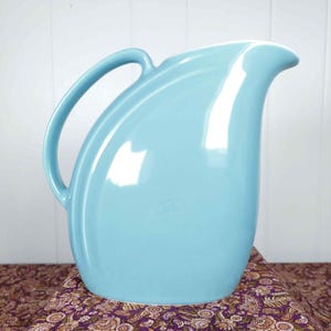 May include: A light blue ceramic pitcher with a curved handle and a spout. The pitcher has a glossy finish and sits on a patterned fabric. The pitcher is a vintage style and is suitable for serving beverages.