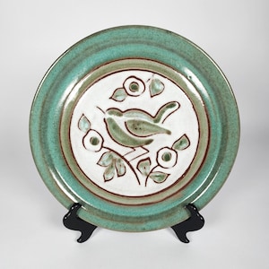 May include: A ceramic plate with a teal rim and a white center featuring a green bird perched on a branch with leaves and flowers. The plate has a brown border and is displayed on a black stand. The plate is a decorative item.