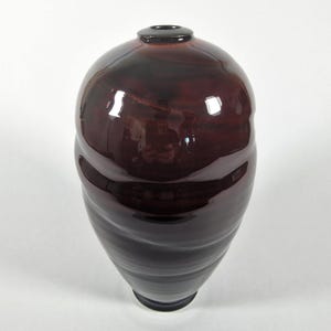 May include: A glossy, dark burgundy ceramic vase with a bulbous top and a tapered, ridged base. The vase has a small, dark opening at the top and reflects light, creating highlights. The overall shape is organic and elegant, suitable for home decor.