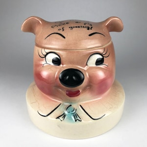 May include: A vintage ceramic pig-shaped cookie jar with a lid. The pig has a pink face, large eyes, and a black nose. The jar has a white collar with a blue bow and the words "Make a pig of yourself!" written on the lid.