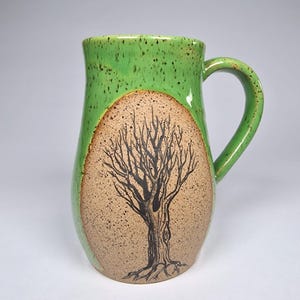 May include: A handmade ceramic mug with a green speckled glaze and a brown, textured oval featuring a black tree design. The mug has a curved handle and a wide opening, perfect for coffee or tea.