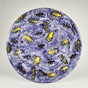 Puede incluir: Un plato redondo de cerámica con un diseño pintado a mano con un fondo azul con pinceladas. El plato está decorado con insectos amarillos y negros. Los insectos son de varios tamaños y posiciones, creando un patrón caprichoso.