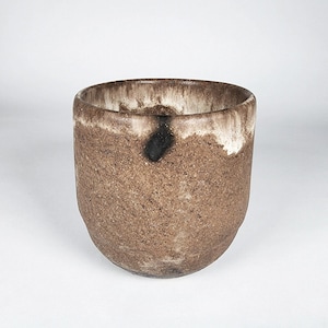 May include: A handcrafted, cylindrical ceramic cup with a textured, speckled brown exterior. The rim features a gradient of white and brown, with a dark black spot on the side. The cup has a rounded base and is set against a white background.