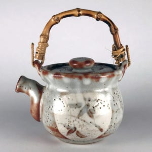 Può includere: Una teiera in ceramica con manico in bambù. La teiera ha un corpo grigio chiaro e bianco con dettagli marroni e un coperchio abbinato. Il manico è in bambù curvo ed è fissato con spago intrecciato.