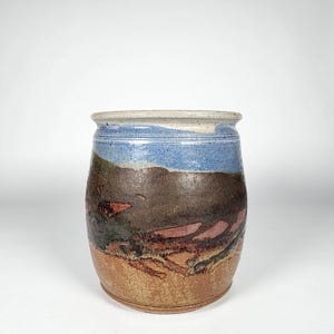 Puede incluir: Un jarrón de cerámica hecho a mano con un diseño de paisaje. El jarrón presenta una banda azul claro en la parte superior, una sección media marrón y roja y una base beige. La cerámica tiene un aspecto rústico y texturizado.