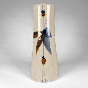 May include: A tall, cylindrical ceramic vase with a smooth, off-white glazed finish. The surface features a vertical design consisting of two small, dark brown circular accents and a central abstract leaf motif painted in shades of blue and brown.
