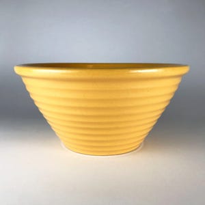 May include: A yellow ceramic mixing bowl with a ribbed exterior. The bowl has a wide rim and a rounded, conical shape. The color is a bright, sunny yellow, and the bowl appears to be in good condition.
