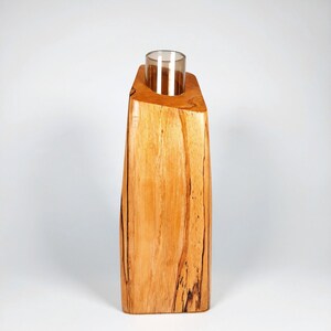 Op de afbeelding: Een houten kandelaar met een heldere glazen inzet. De houder is gemaakt van lichtgekleurd hout met donkere nerfpatronen. De bovenkant is schuin en de glazen inzet is cilindrisch.