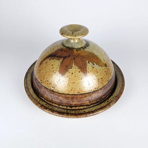 Puede incluir: Una mantequera de cerámica con tapa abovedada y base circular. La tapa presenta un diseño floral marrón y un pequeño asa. El plato tiene una paleta de colores moteados y terrosos con tonos marrones y beige, creando una estética rústica.