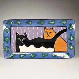Puede incluir: Bandeja rectangular de cerámica con un borde azul con diseños de hojas y flechas verdes. El centro de la bandeja tiene un fondo morado con líneas onduladas blancas y un gato negro con ojos verdes y un gato naranja.