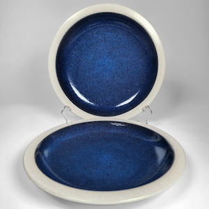 Puede incluir: Dos platos redondos de cerámica con un centro esmaltado en azul profundo moteado y un borde ancho de color blanco roto mate. Los platos tienen un acabado liso y brillante en el centro y una textura contrastante en el borde exterior.
