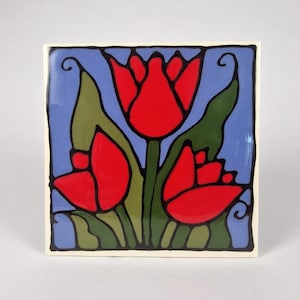 Könnte beinhalten: Quadratische Keramikfliese mit stilisiertem Blumendesign. Sie zeigt drei rote Tulpen mit grünen Blättern auf hellblauem Hintergrund. Das Design ist schwarz umrandet, mit einem cremefarbenen Rand.