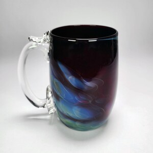 May include: A handmade glass mug with a clear handle and a dark burgundy body. The mug features swirling blue, green, and purple accents. The handle is attached with decorative glass elements.