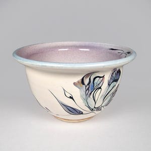 May include: A ceramic bowl with a cream-colored exterior and a lavender interior. The rim is outlined in light blue. The bowl is decorated with a floral design in shades of blue, purple, and black.