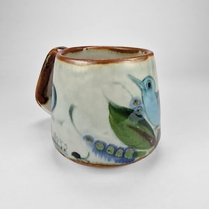 May include: A ceramic mug with a white base, featuring a blue bird and green leaf design. The mug has a brown handle and rim, with a slightly textured surface. The mug is likely handmade and suitable for hot beverages.