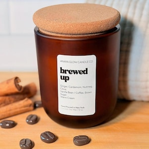May include: A brown glass candle jar with a cork lid, labeled "brewed up." The label lists ingredients like ginger, cardamom, and vanilla bean. Cinnamon sticks and coffee bean-shaped candies are scattered around the candle on a wooden surface. The candle is 12 oz.