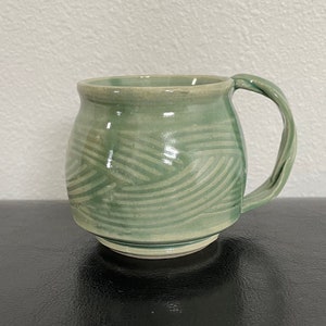 May include: A green ceramic mug with a white glaze and a wavy pattern. The mug has a rounded body and a handle.