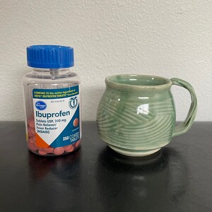 May include: A bottle of Kroger brand Ibuprofen tablets, 200 mg, with a blue lid and pink pills inside. The bottle is sitting on a black surface next to a green ceramic mug with a white handle.