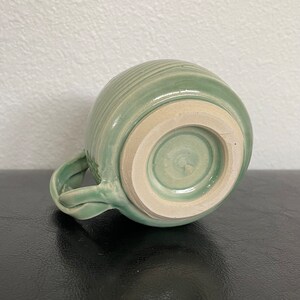 May include: A light green ceramic mug with a textured, ridged exterior and a white glazed interior. The mug has a handle and a rounded base.