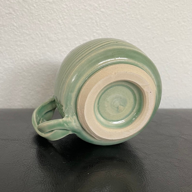 May include: A light green ceramic mug with a textured, ridged exterior and a white glazed interior. The mug has a handle and a rounded base.