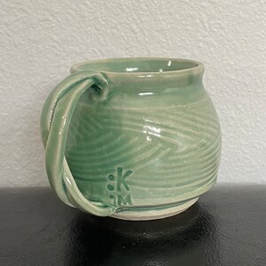 May include: A green ceramic mug with a unique, twisted handle and a subtle, wavy pattern. The mug is signed with the initials "SKM" on the bottom.