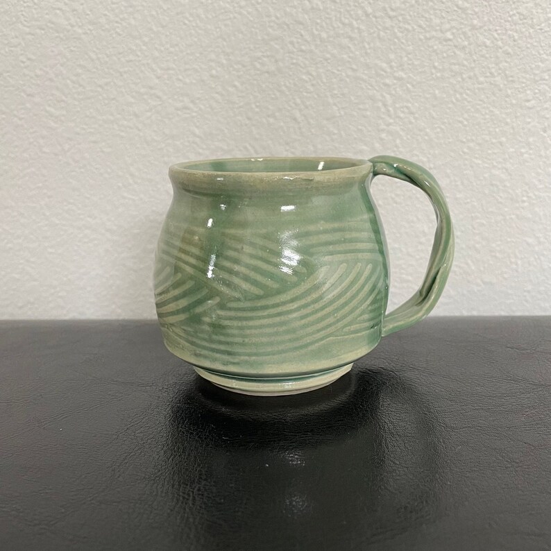 May include: A green ceramic mug with a white glaze and a wavy pattern. The mug has a rounded body and a handle.
